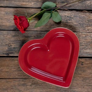 May include: A red heart-shaped ceramic plate on a wooden surface. A single red rose with green leaves is lying on the surface.