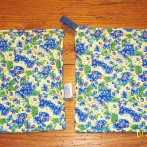Op de afbeelding: Twee vierkante pannenlappen met een blauw en groen bosbessenpatroon op een gele achtergrond. Elke pannenlap heeft een blauwe lus om op te hangen en een klein wit label.