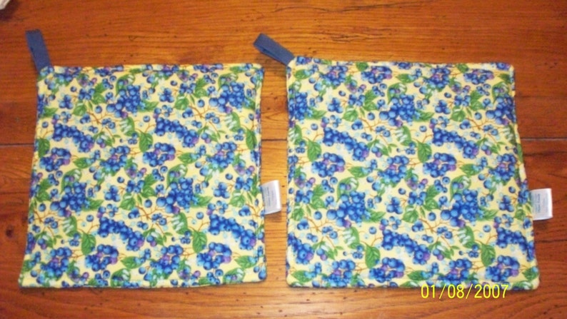 Op de afbeelding: Twee vierkante pannenlappen met een blauw en groen bosbessenpatroon op een gele achtergrond. Elke pannenlap heeft een blauwe lus om op te hangen en een klein wit label.