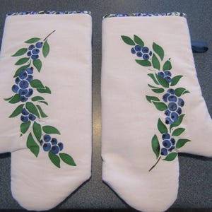 May include: A pair of white oven mitts with a blue and green blueberry design. The mitts have a blue loop for hanging and a blue and green patterned trim. The blueberry design is painted on the front of each mitt.