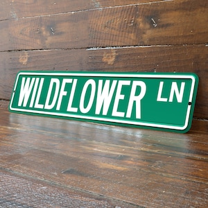 May include: A green metal street sign with the words "WILDFLOWER LN" in white block letters. The sign has a white border and is leaning against a wooden surface.