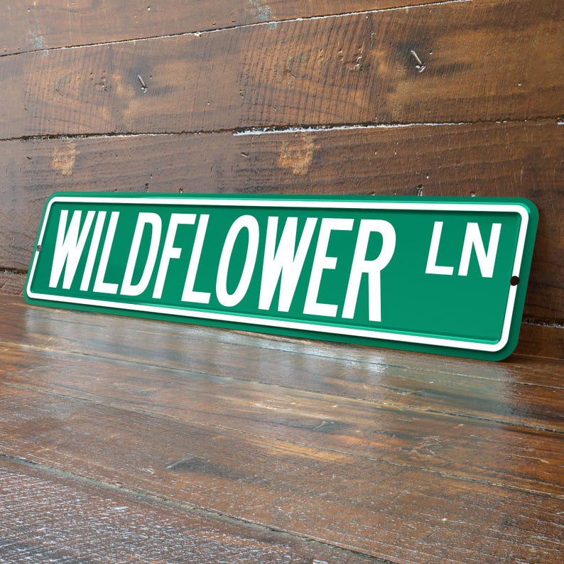 May include: A green metal street sign with the words "WILDFLOWER LN" in white block letters. The sign has a white border and is leaning against a wooden surface.