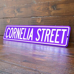 May include: A purple street sign with the words "CORNELIA STREET" in white, set against a wooden background. The sign has a white border and is angled slightly, showcasing its rectangular shape.