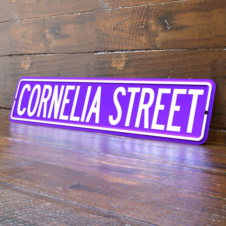 May include: A purple street sign with the words "CORNELIA STREET" in white, set against a wooden background. The sign has a white border and is angled slightly, showcasing its rectangular shape.