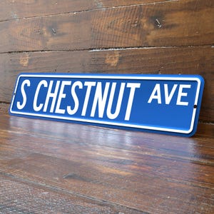May include: A blue street sign with white lettering that reads "S CHESTNUT AVE". The sign has a white border and is mounted on a wooden surface. The sign is angled slightly, showing the text in a clear, readable font.