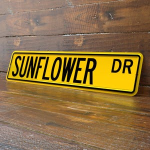 May include: A yellow metal sign with the words "SUNFLOWER DR" in black, set against a wooden background. The sign has a black border and is slightly angled, showcasing its rectangular shape.