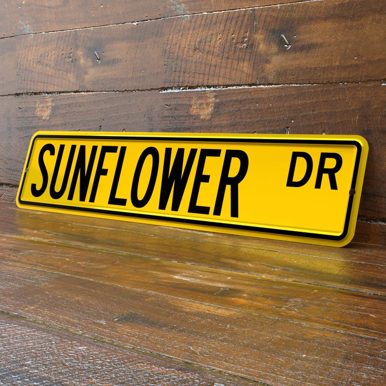 May include: A yellow metal sign with the words "SUNFLOWER DR" in black, set against a wooden background. The sign has a black border and is slightly angled, showcasing its rectangular shape.
