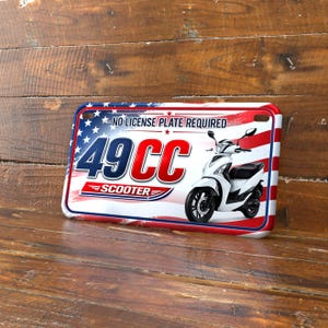 May include: A decorative license plate featuring a white scooter design. The plate has a red, white, and blue American flag theme with the text "NO LICENSE PLATE REQUIRED" and "49CC SCOOTER".