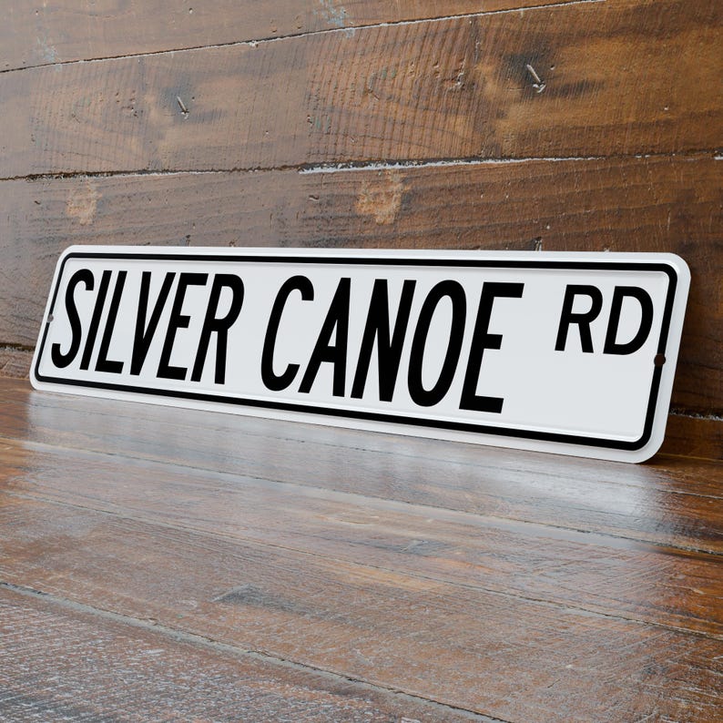 May include: A rectangular white metal sign with the words "SILVER CANOE RD" in bold black letters. The sign has a black border and is angled slightly, resting on a wooden surface.