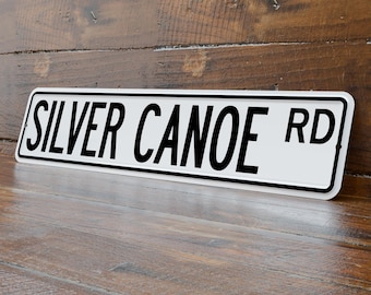 May include: A rectangular white metal sign with the words "SILVER CANOE RD" in bold black letters. The sign has a black border and is angled slightly, resting on a wooden surface.