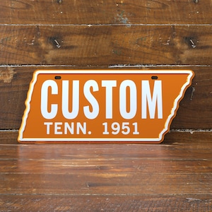 Könnte beinhalten: Orangefarbenes Nummernschild mit weißem Text "CUSTOM" über "TENN. 1951". Das Schild hat die Form des Bundesstaates Tennessee. Der Hintergrund ist eine Holzoberfläche.