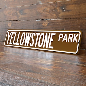 May include: A rectangular brown metal sign with the words "YELLOWSTONE PARK" in white, outlined in a white border. The sign is angled against a wooden background.