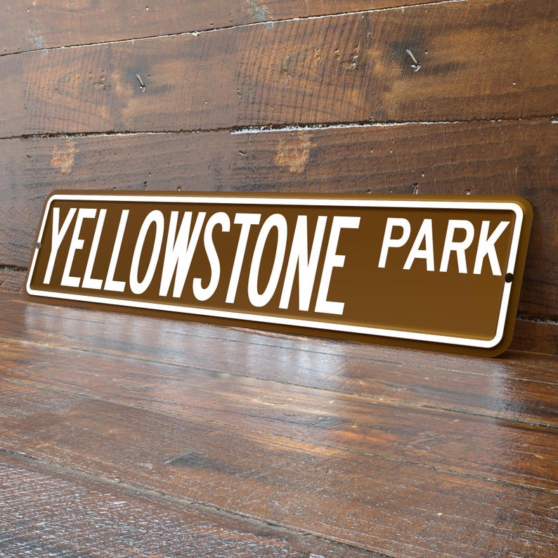 May include: A rectangular brown metal sign with the words "YELLOWSTONE PARK" in white, outlined in a white border. The sign is angled against a wooden background.