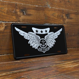 May include: Black license plate with a silver emblem featuring wings, three stars, an anchor, and the words "AIR CREW." The plate is rectangular with rounded corners and mounting holes.