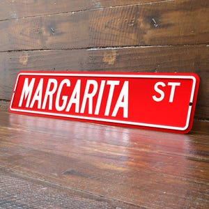 May include: A red street sign with white lettering that reads "MARGARITA ST". The sign has a white border and is mounted on a wooden surface. The sign is angled slightly, giving a three-dimensional appearance.