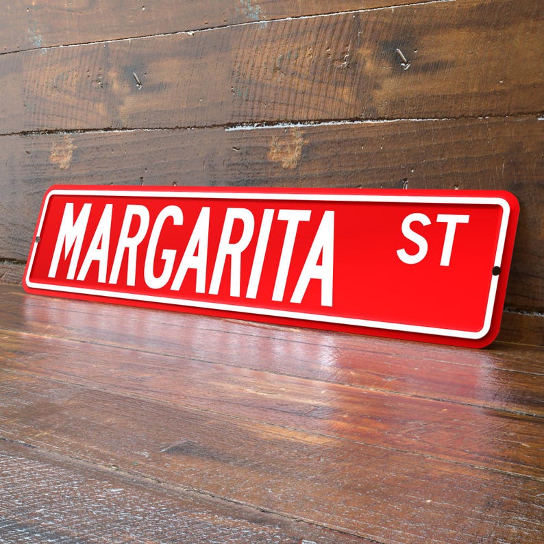 May include: A red street sign with white lettering that reads "MARGARITA ST". The sign has a white border and is mounted on a wooden surface. The sign is angled slightly, giving a three-dimensional appearance.