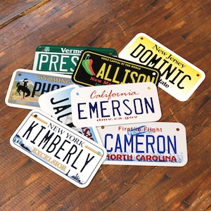 May include: A collection of novelty license plates with various state names and personalized names. The plates feature designs from states like New York, California, and Wyoming, with names such as Kimberly, Emerson, and Dominic. The plates are arranged on a wooden surface.