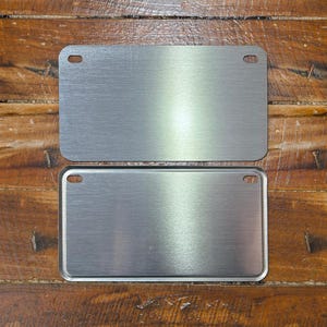 May include: Two blank metal license plates with rounded corners and holes for hanging. The plates are silver and have a brushed finish.