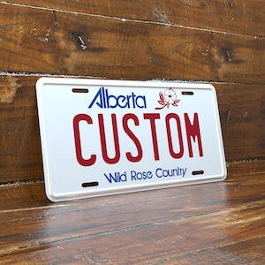 Peut inclure: Plaque d'immatriculation blanche avec le mot "Alberta" en bleu, "CUSTOM" en rouge et "Wild Rose Country" en bleu. Un emblème floral rouge est au-dessus du mot "CUSTOM". Sur un fond en bois.