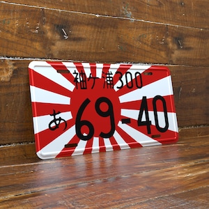 May include: A rectangular license plate with a red sunburst design and white rays. The plate features Japanese characters and the numbers "69-40" in black. The background is a wooden surface.