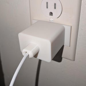 May include: A white power adapter plugged into a wall outlet. The adapter is square and has a white cord extending from it. The wall outlet is off-white and has two vertical slots and a circular opening.