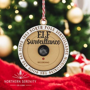 以下が含まれることがあります： 白い背景と茶色の文字で書かれた丸い木製のクリスマスオーナメント。テキストには「エルフ監視」と赤い点、その下に「録音中」と書いてあります。オーナメントには、縁に「善と悪は1863年から北極監視監視」という文字も書かれています。