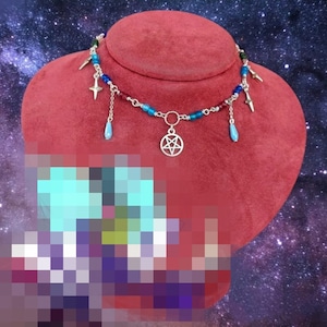 May include: A silver-toned choker necklace with a pentagram pendant and dangling charms. The necklace features blue, red, and green beads, with silver crosses and teardrop-shaped charms. The necklace is displayed on a red velvet bust against a starry background.