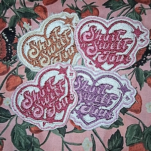 May include: Four heart-shaped embroidered patches with the text "Shut Sweet You" in various colors: orange, pink, red, and purple. The patches are on a pink fabric with a strawberry and butterfly pattern.