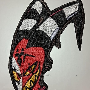 Può includere: Toppa ricamata raffigurante un personaggio dei cartoni animati rosso, nero e bianco con denti e corna gialle. La toppa ha un bordo nero ed è progettata per essere stirata o cucita su tessuto. Il design è dettagliato e colorato.