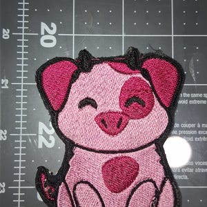 Könnte beinhalten: Gestickter Aufnäher mit einem lächelnden Schwein mit rosa und dunkelrosa Details. Das Schwein hat einen großen Fleck auf der Seite und ist schwarz umrandet. Der Aufnäher ist etwa 5 cm breit.