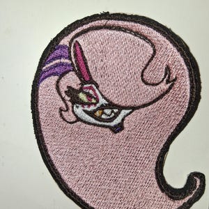May include: A pink embroidered patch depicting a cartoon character with a menacing grin. The character has purple and pink details, with a black border. The patch is circular with a curved tail-like shape.