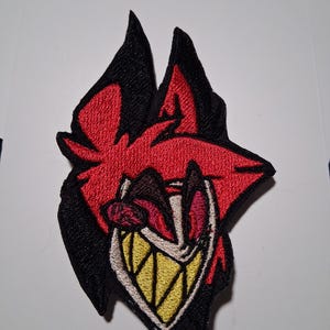 May include: Embroidered patch depicting a stylized red and black character with a wide, toothy grin. The patch has a detailed design with yellow teeth, a white mouth, and red accents. Suitable for clothing or accessories.