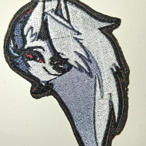 Pode incluir: Patch bordado com a cabeça estilizada de um lobo com pelo branco, cinza e azul. O lobo tem olhos vermelhos e nariz e boca pretos. O patch tem uma borda preta.