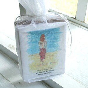 May include: A white gift bag with a white sheer fabric tied around it. The bag has a cartoon drawing of a woman in a bikini on a beach. The text on the bag reads "This is what we call holiday wrapping. Happy Holidays!"