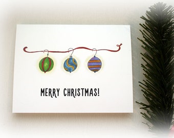 May include: A white card with a red ribbon and three colorful ornaments hanging from it. The text "Merry Christmas!" is printed below the ornaments.