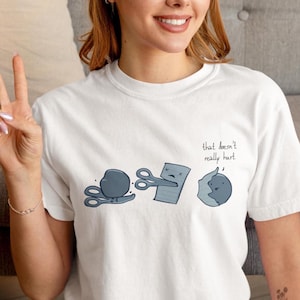 Może przedstawiać: Biała koszulka z zabawną ilustracją kamień, papier, nożyce. Projekt przedstawia kamień, nożyczki i papier z napisem "that doesn't really hurt". Projekt jest w odcieniach szarości i czerni.