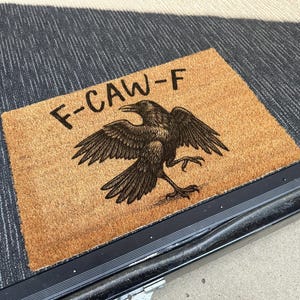 Op de afbeelding: Een rechthoekige deurmat met een bruin kokosoppervlak. De mat heeft een zwarte illustratie van een raaf met uitgespreide vleugels en de tekst "F-CAW-F" in zwarte, gestileerde letters. De mat ligt op een donkergrijze en zwarte textuur.