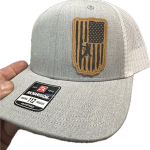 Può includere: Cappellino da camionista grigio e bianco con una toppa in pelle marrone che presenta una bandiera americana con la silhouette di una persona che sale su una scala. La toppa ha un bordo cucito. Il cappello ha un'etichetta nera con il testo "Richardson" e "112 Trucker" in bianco.
