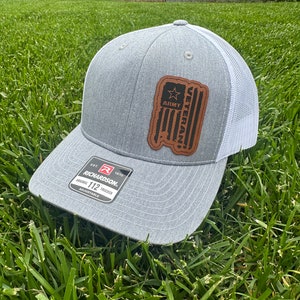 Könnte beinhalten: Eine graue und weiße Trucker-Kappe mit einem braunen Leder-Patch, auf dem "Army Veteran" mit einem Stern und einem amerikanischen Flaggen-Design steht. Die Kappe ist von Richardson 112.