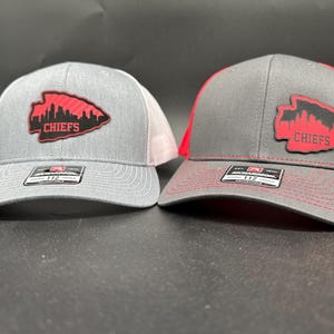 Puede incluir: Dos gorras de camionero grises y rojas con un parche de cuero negro que presenta un contorno rojo del horizonte de Kansas City y la palabra "CHIEFS".