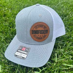 May include: A grey and white trucker hat with a brown leather patch that says "Let's Keep The Dumbfuckery To A Minimum Today". The hat is made by Richardson and is adjustable.