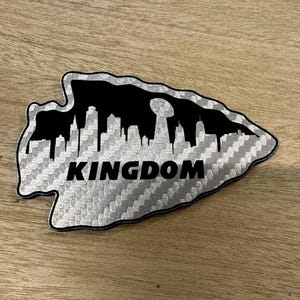 May include: A silver and black sticker in the shape of an arrowhead. The sticker features a cityscape silhouette in black, with the word "KINGDOM" in bold black letters. The background has a textured, carbon fibre-like pattern.