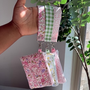 May include: A collection of floral and gingham patterned lanyards with matching zippered pouches. The lanyards feature pink, green, and white designs, with silver-tone hardware. The pouches are in various floral patterns and colors.