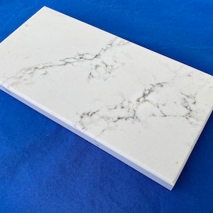 May include: A white countertop sample with a gray veining pattern. The sample is rectangular and has a smooth, polished surface.