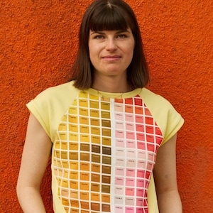 Könnte beinhalten: Eine Frau trägt ein gelbes, kurzärmeliges Shirt mit einem bunten Rastermuster und blaue Jeans. Das Shirt hat einen runden Ausschnitt und Raglanärmel.