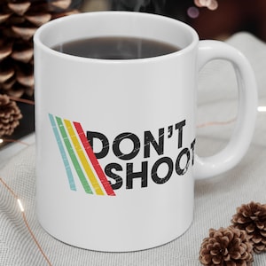 May include: White ceramic mug with the text "DON'T SHOOT" in black, with a colorful striped design. The mug is filled with a dark liquid and sits on a light gray surface with decorative elements.