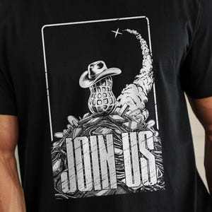 Puede incluir: Camiseta negra con un gráfico de un cacahuete con sombrero de vaquero y las palabras "JOIN US". El diseño incluye una estela de cohete estilizada. La palabra "BLACK" está en la parte inferior.