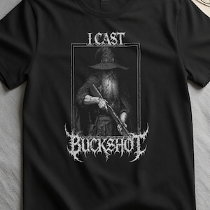 May include: A black short-sleeved t-shirt featuring a white graphic print of a wizard holding a shotgun. The text "I CAST" appears above the wizard, and the word "BUCKSHOT" is printed below in a stylized, jagged font within a rectangular frame.