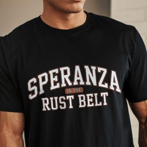 Könnte beinhalten: Schwarzes T-Shirt mit Rundhalsausschnitt und dem Text "SPERANZA in the RUST BELT" in weißer und brauner Schrift. Das Wort "BLACK" ist in einem schwarzen Oval am unteren Bildrand aufgedruckt. Das Shirt ist aus weichem Material.