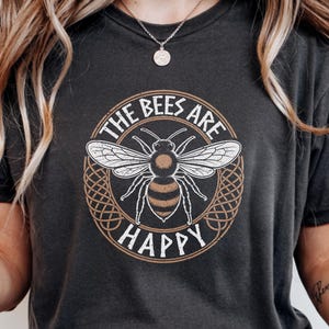 Może przedstawiać: Czarna koszulka z melanżem z okrągłą grafiką przedstawiającą pszczołę i napis "THE BEES ARE HAPPY". Pszczoła jest biało-brązowa, wewnątrz brązowo-białej dekoracyjnej obwódki. Koszulka jest w ciemnoszarym kolorze.